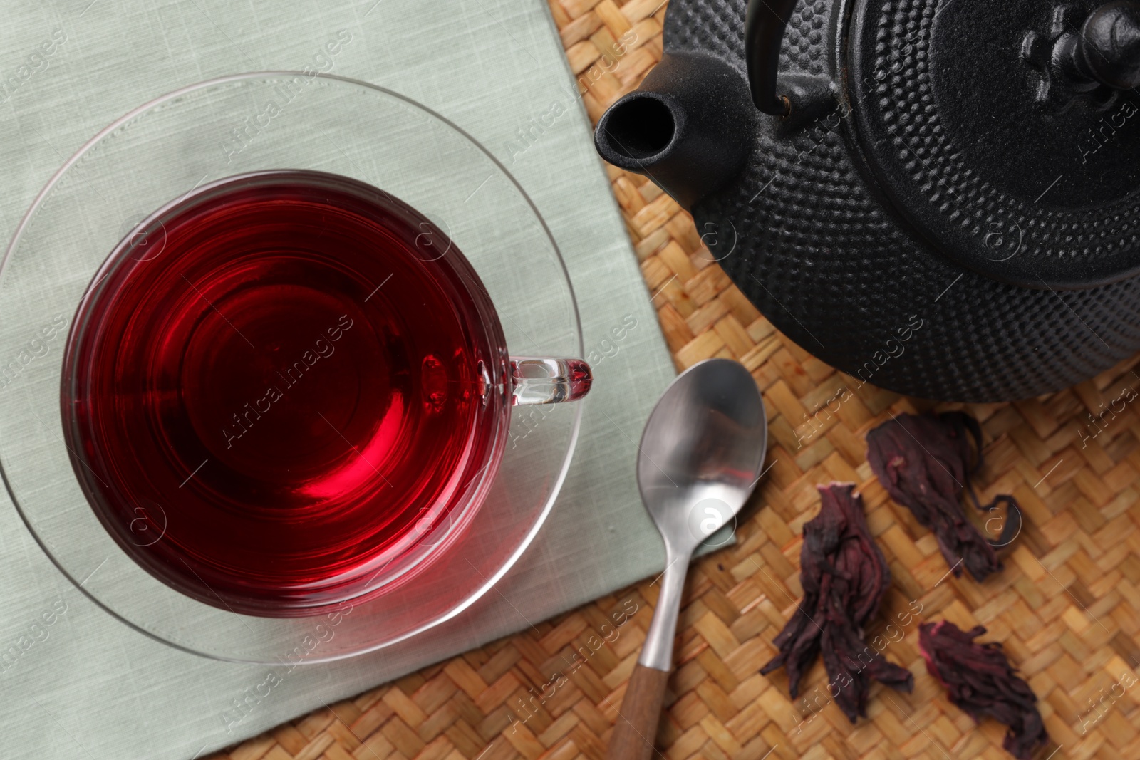 Delicious hibiscus tea in glass cup, teapot, dry roselle sepals and spoon on wicker mat, flat lay Photo of Delicious hibiscus tea in glass cup, teapot, dry roselle sepals and spoon on wicker mat, flat lay