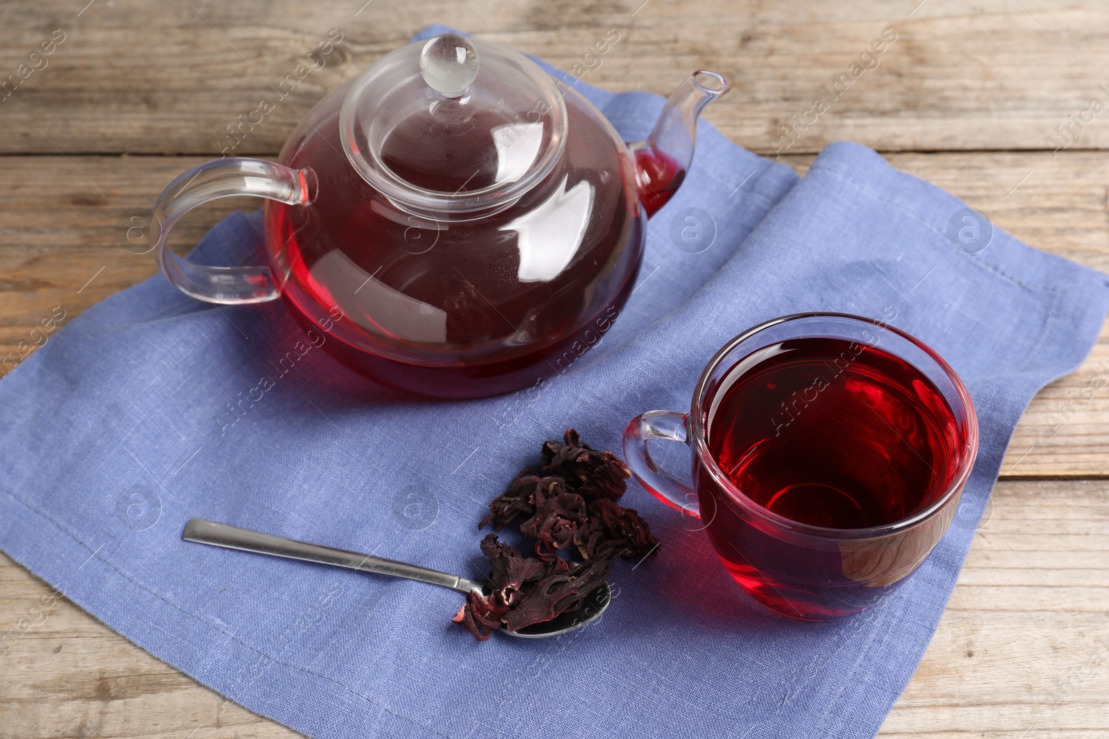 Delicious hibiscus tea in glass cup, teapot, dry roselle sepals and spoon on wooden table Photo of Delicious hibiscus tea in glass cup, teapot, dry roselle sepals and spoon on wooden table