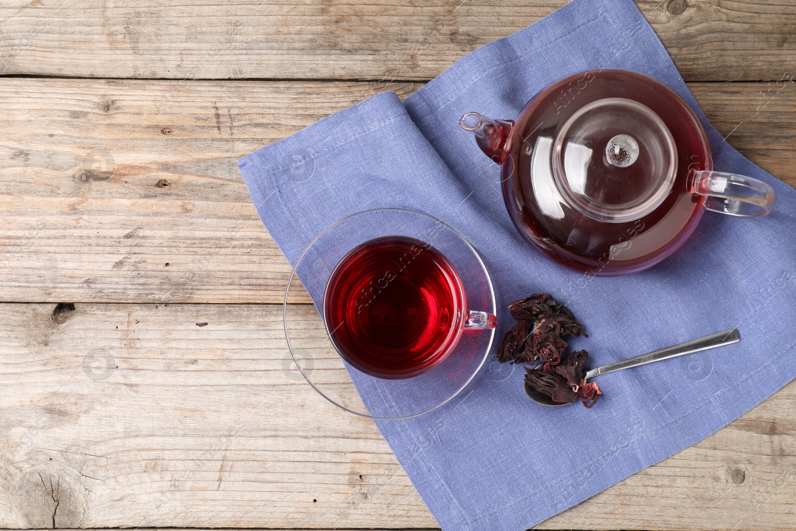 Delicious hibiscus tea in glass cup, teapot, dry roselle sepals and spoon on wooden table, flat lay. Space for text Photo of Delicious hibiscus tea in glass cup, teapot, dry roselle sepals and spoon on wooden table, flat lay. Space for text