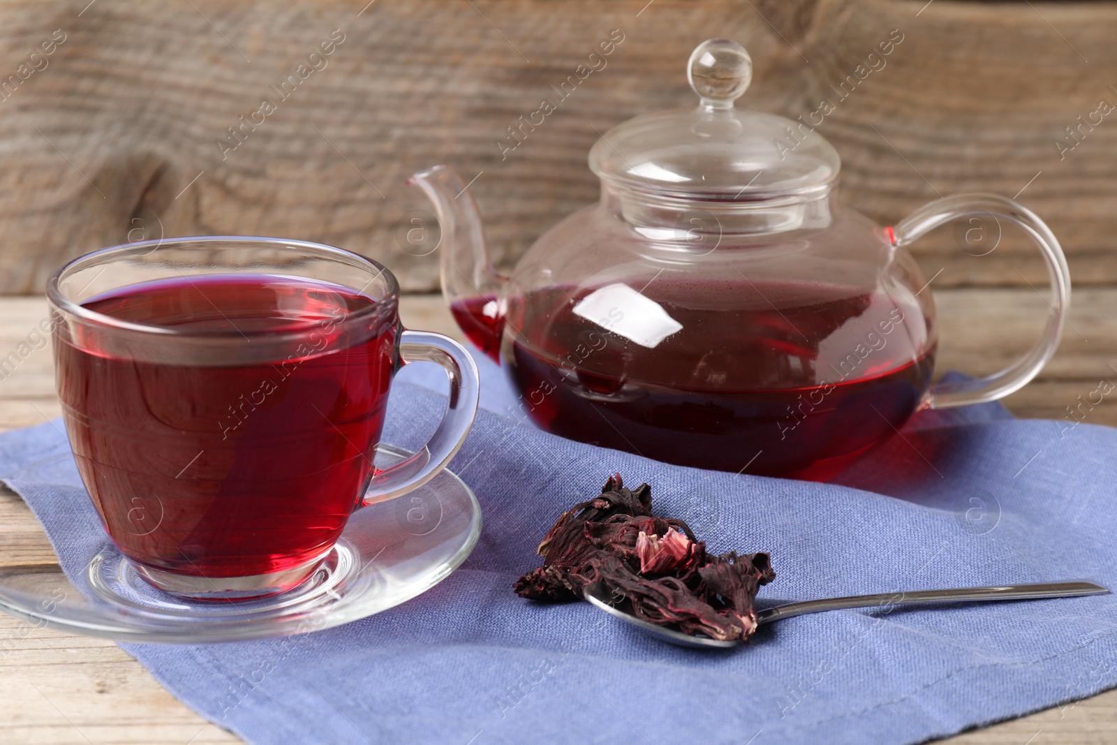Delicious hibiscus tea in glass cup, teapot, dry roselle sepals and spoon on wooden table, closeup Photo of Delicious hibiscus tea in glass cup, teapot, dry roselle sepals and spoon on wooden table, closeup