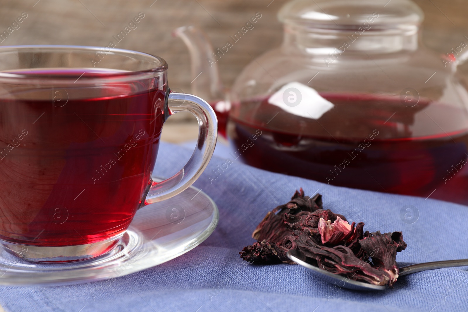 Delicious hibiscus tea in glass cup, teapot, dry roselle sepals and spoon on table, closeup Photo of Delicious hibiscus tea in glass cup, teapot, dry roselle sepals and spoon on table, closeup