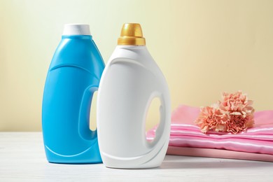 Photo of Bottles of fabric softener, clean clothes and carnation flowers on white wooden table against light yellow background