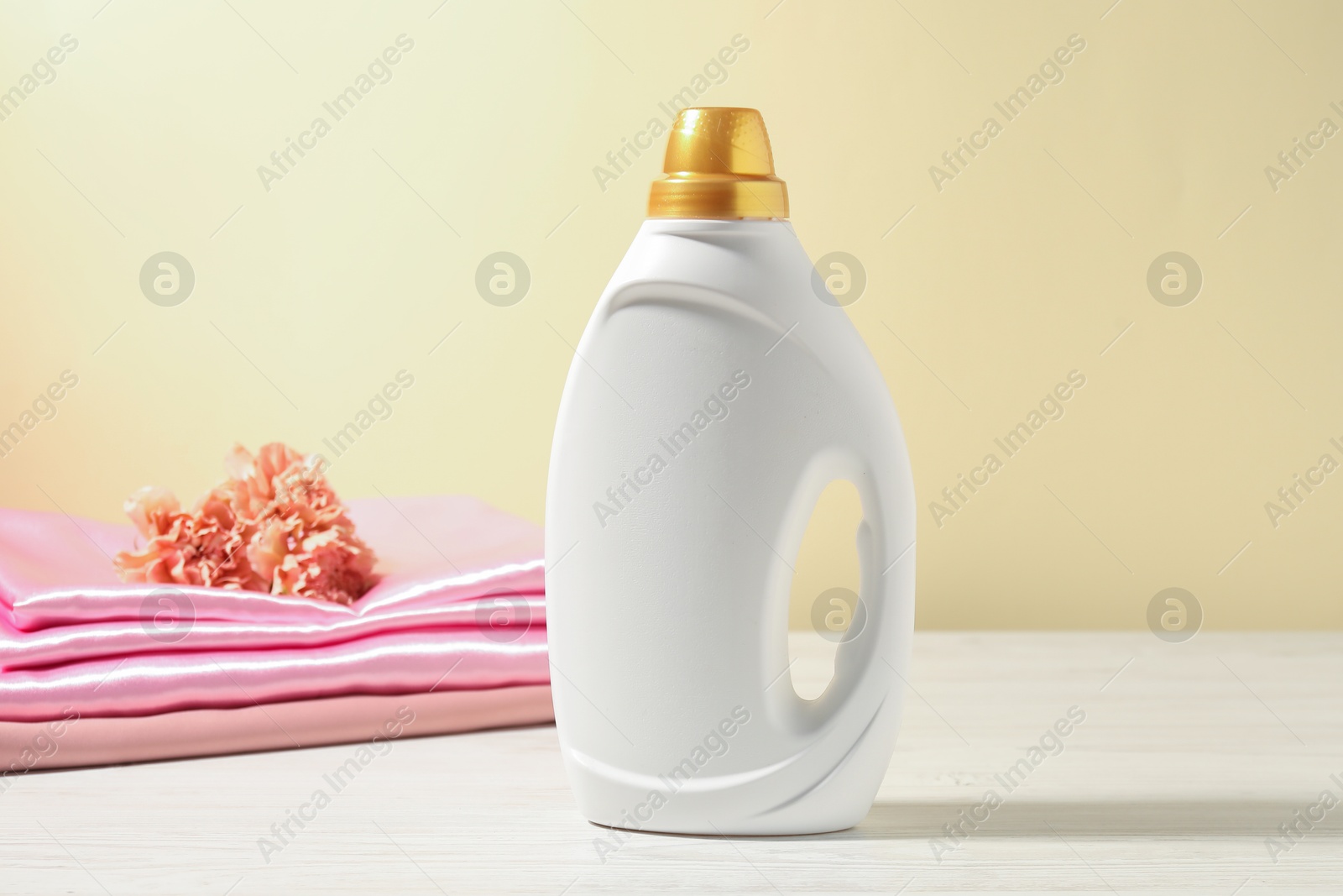 Bottle of fabric softener, clean clothes and carnation flowers on white wooden table against light yellow background Photo of Bottle of fabric softener, clean clothes and carnation flowers on white wooden table against light yellow background
