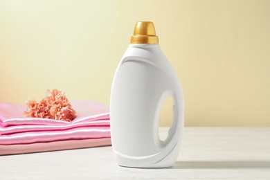 Photo of Bottle of fabric softener, clean clothes and carnation flowers on white wooden table against light yellow background