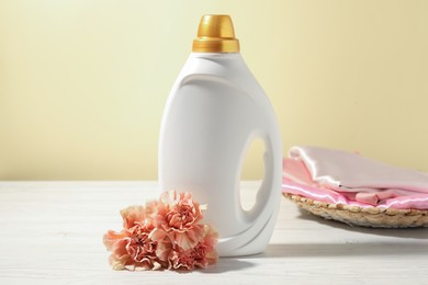 Photo of Bottle of fabric softener, clean clothes and carnation flowers on white wooden table against light yellow background