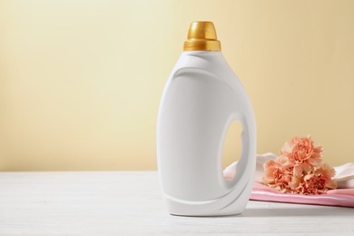 Photo of Bottle of fabric softener, clean clothes and carnation flowers on white wooden table against light yellow background. Space for text