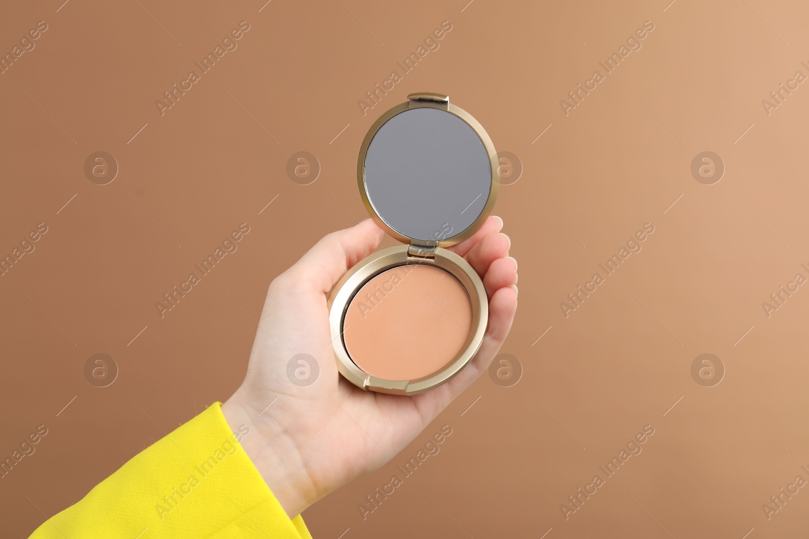Photo of Woman holding open face powder with mirror on brown background, closeup