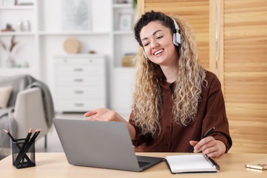 Photo of Smiling woman in headphones having videochat by laptop at desk in home office
