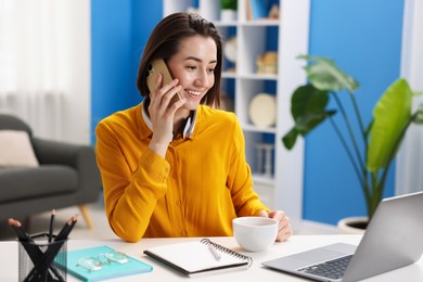 Photo of Smiling woman with laptop talking by smartphone at desk in home office