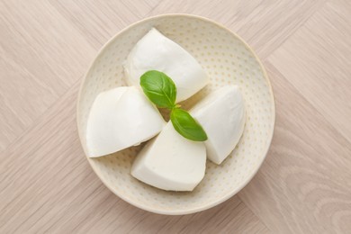 Photo of Pieces of delicious mozzarella cheese and basil in bowl on wooden table, top view