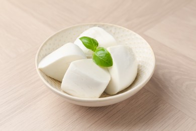 Photo of Pieces of delicious mozzarella cheese and basil in bowl on wooden table, closeup