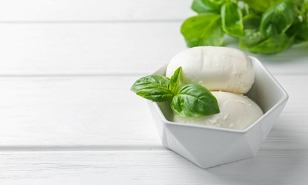 Photo of Pieces of delicious mozzarella cheese and basil in bowl on white wooden table, closeup. Space for text