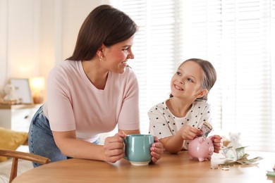 Photo of Mother and daughter putting pocket money into piggybank at table indoors