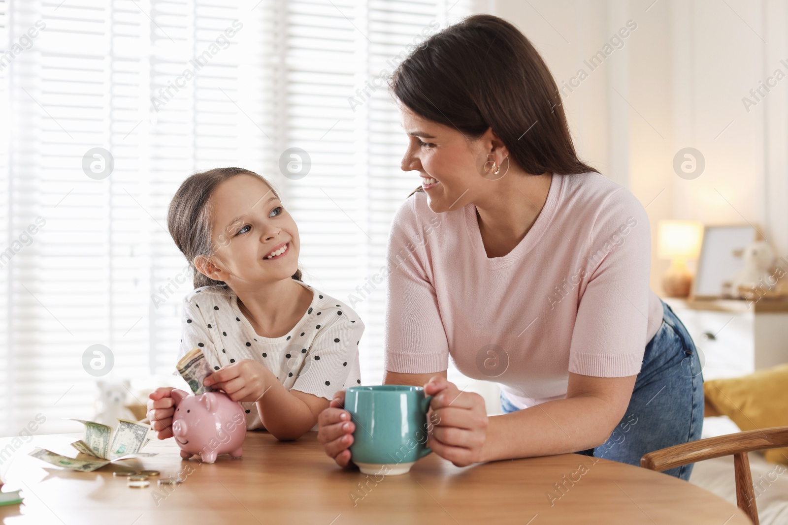 Mother and daughter putting pocket money into piggybank at table indoors Photo of Mother and daughter putting pocket money into piggybank at table indoors