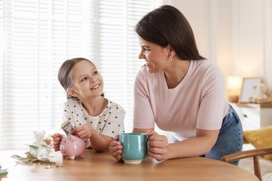 Photo of Mother and daughter putting pocket money into piggybank at table indoors