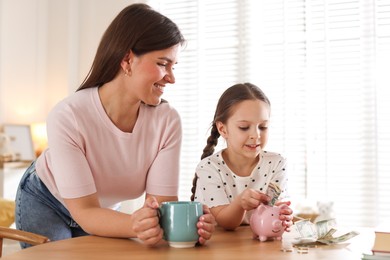 Photo of Mother and daughter putting pocket money into piggybank at table indoors
