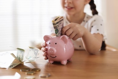 Photo of Girl putting pocket money into piggybank at table indoors, closeup