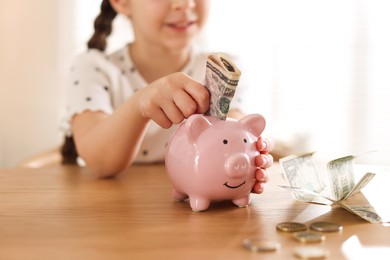 Photo of Girl putting pocket money into piggybank at table indoors, closeup
