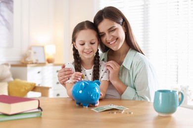 Photo of Mother and daughter putting pocket money into piggybank at table indoors