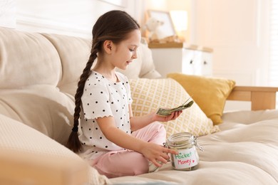 Photo of Girl putting her pocket money into jar on sofa at home