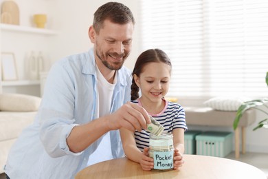 Photo of Father and daughter putting pocket money into jar at table indoors