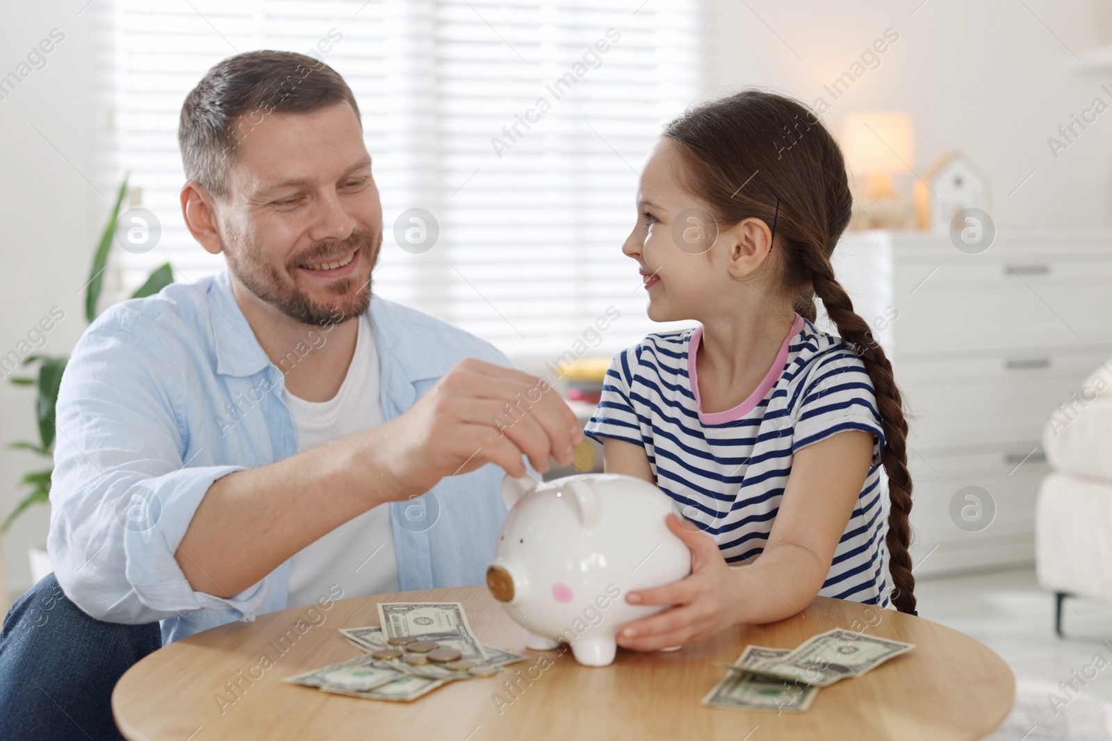 Father and daughter putting pocket money into piggybank at table indoors Photo of Father and daughter putting pocket money into piggybank at table indoors