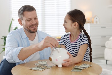Photo of Father and daughter putting pocket money into piggybank at table indoors