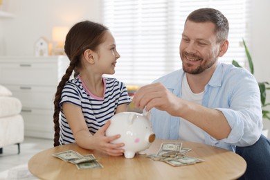 Photo of Father and daughter putting pocket money into piggybank at table indoors