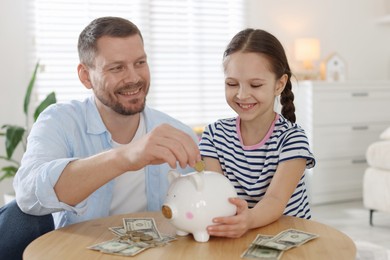 Photo of Father and daughter putting pocket money into piggybank at table indoors