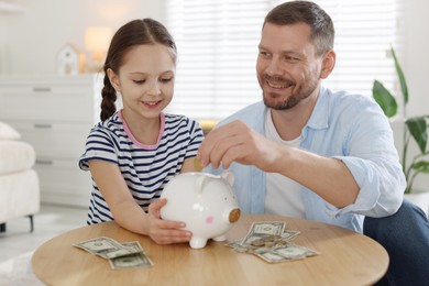 Photo of Father and daughter putting pocket money into piggybank at table indoors