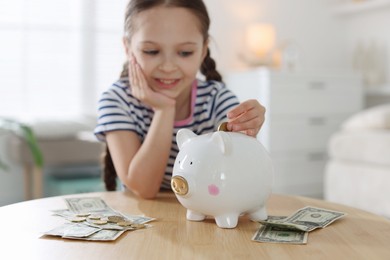 Photo of Girl putting her pocket money into piggybank at table indoors, selective focus
