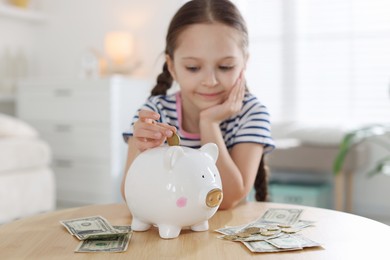 Photo of Girl putting her pocket money into piggybank at table indoors, selective focus