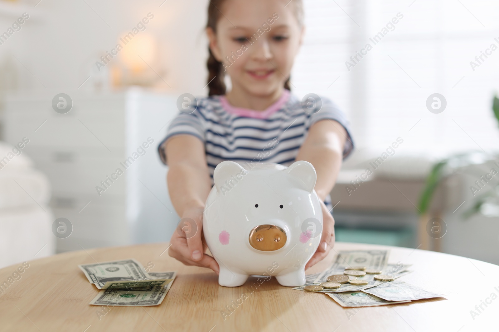 Girl with piggybank and pocket money at table indoors, selective focus Photo of Girl with piggybank and pocket money at table indoors, selective focus