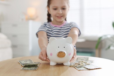 Photo of Girl with piggybank and pocket money at table indoors, selective focus