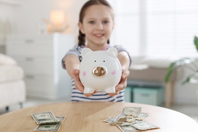 Photo of Girl with piggybank and pocket money at table indoors, selective focus
