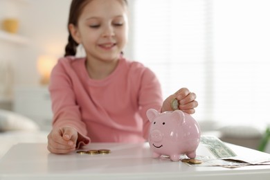 Photo of Girl putting her pocket money into piggybank at table indoors, selective focus