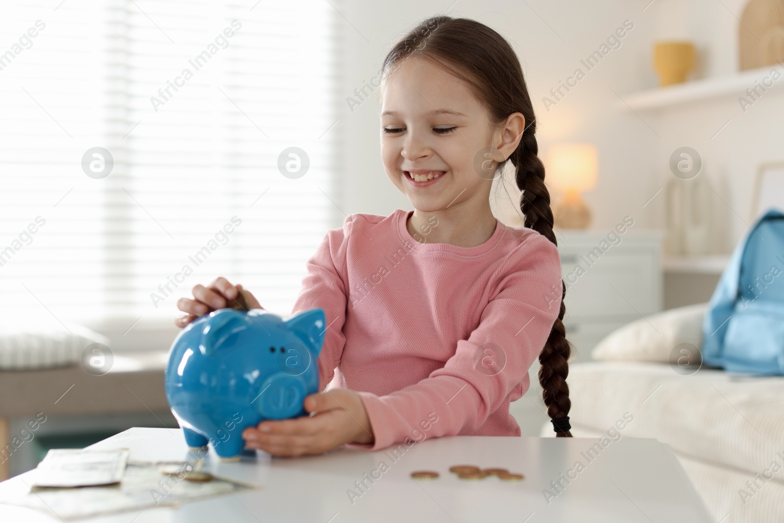 Girl putting her pocket money into piggybank at table indoors Photo of Girl putting her pocket money into piggybank at table indoors