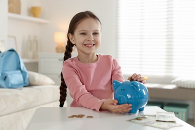 Photo of Girl putting her pocket money into piggybank at table indoors