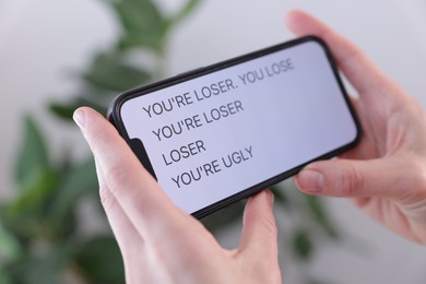 Photo of Cyber bullying. Woman holding smartphone with insults on blurred background, closeup