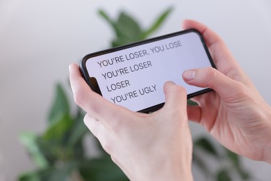 Photo of Cyber bullying. Woman holding smartphone with insults on blurred background, closeup