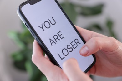 Photo of Cyber bullying. Woman holding smartphone with text You Are Loser on blurred background, closeup