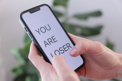 Photo of Cyber bullying. Woman holding smartphone with text You Are Loser on blurred background, closeup