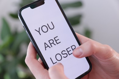 Photo of Cyber bullying. Woman holding smartphone with text You Are Loser on blurred background, closeup