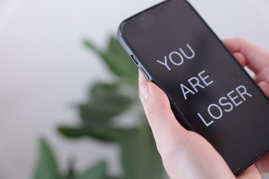 Photo of Cyber bullying. Woman holding smartphone with text You Are Loser on blurred background, closeup. Space for text