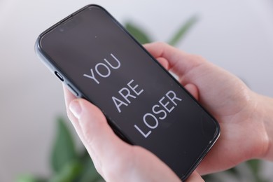 Photo of Cyber bullying. Woman holding smartphone with text You Are Loser on blurred background, closeup