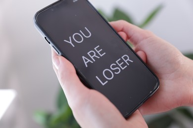 Photo of Cyber bullying. Woman holding smartphone with text You Are Loser on blurred background, closeup