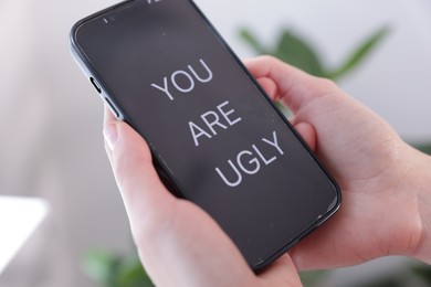 Photo of Cyber bullying. Woman holding smartphone with text You Are Ugly on blurred background, closeup