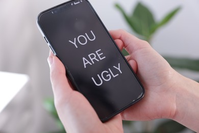 Photo of Cyber bullying. Woman holding smartphone with text You Are Ugly on blurred background, closeup