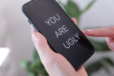 Photo of Cyber bullying. Woman holding smartphone with text You Are Ugly on blurred background, closeup