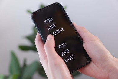 Photo of Cyber bullying. Woman holding smartphone with insults on blurred background, closeup